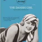Book cover of The Danish Girl by David Ebershoff, featuring a smiling woman in vintage clothing and headscarf, lying down. The cover is light blue with award logos and a gold label noting its soon to be a major motion picture.