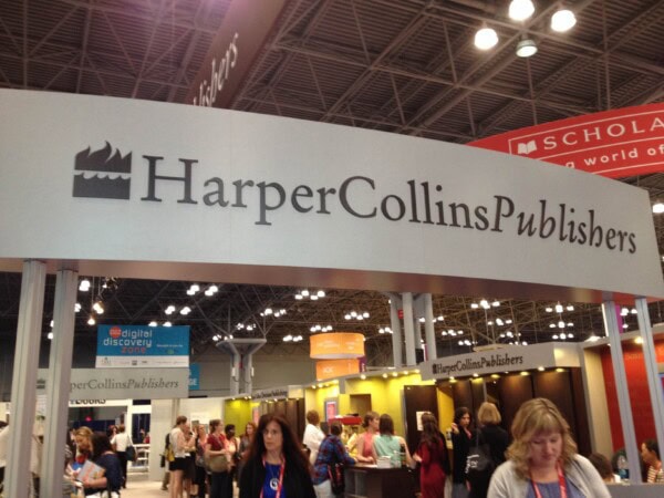 A large HarperCollins Publishers sign hangs above a busy booth at a convention center, with people walking and gathering below. Bright lights and other exhibitor booths are visible in the background.