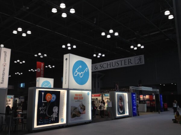 A book fair or convention hall with illuminated booths displaying large book covers, including “Leo: A Ghost Story” and “You Need More Sleep,” under a high ceiling with bright lights and a Simon & Schuster banner.