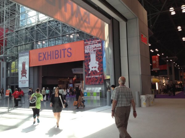 Large convention center hall with high glass ceilings, banners reading EXHIBITS and THE SCORPION RULES, and people walking toward exhibit areas. Bright sunlight fills the space.