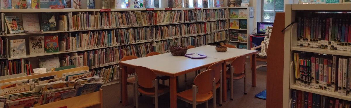 A library children's section with shelves of books, a white table surrounded by small wooden chairs, and a computer in the background.