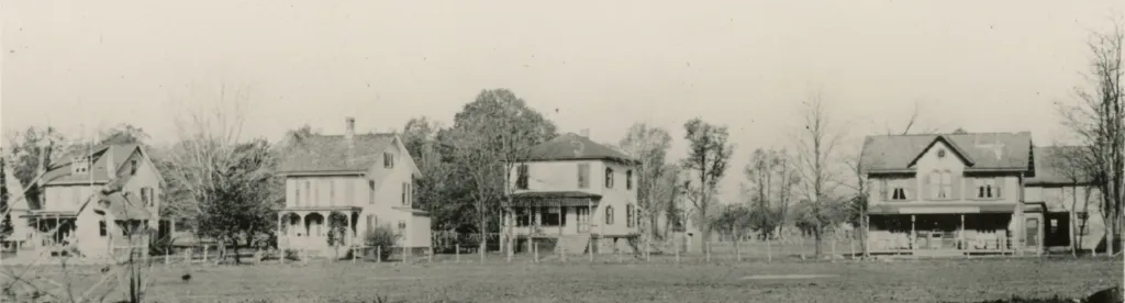 A row of five two-story houses with porches, spaced apart and surrounded by trees and open grassy land.